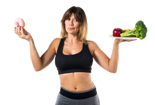 Спортивная женщина держит фрукты и овощи в одной руке и пончик в другой руке — стоковое фото