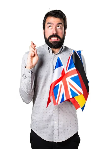 Hombre guapo con barba sosteniendo muchas banderas y con los dedos cruzados —  Fotos de Stock