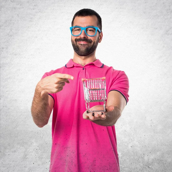 Homme avec des vêtements colorés tenant un jouet de chariot supermarché sur gris — Photo