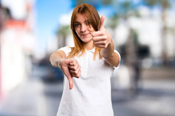 Beautiful young girl making good-bad sign on unfocused background — Stock Photo, Image