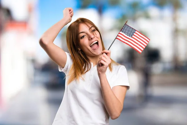 Feliz hermosa joven sosteniendo una bandera americana sobre fondo desenfocado —  Fotos de Stock