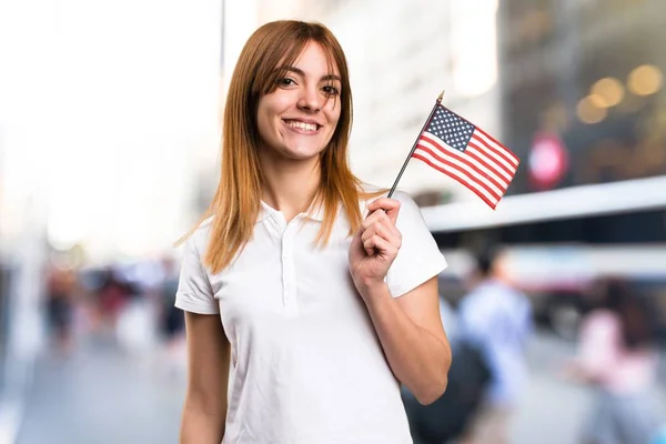 Feliz hermosa joven sosteniendo una bandera americana sobre fondo desenfocado —  Fotos de Stock