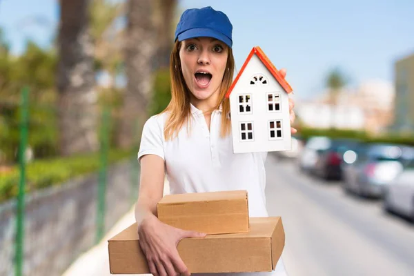 Surprised Delivery woman holding a little house on unfocused background — Stock Photo, Image