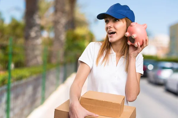 Surprised Delivery woman holding a piggybank on unfocused background