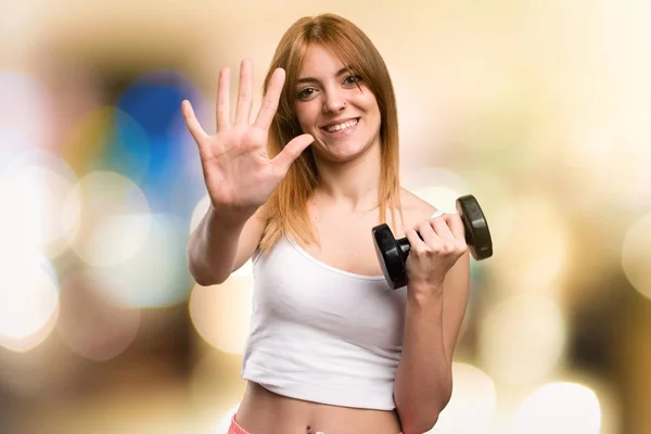 Hermosa mujer deportiva con mancuernas contando cinco sobre fondo desenfocado — Foto de Stock