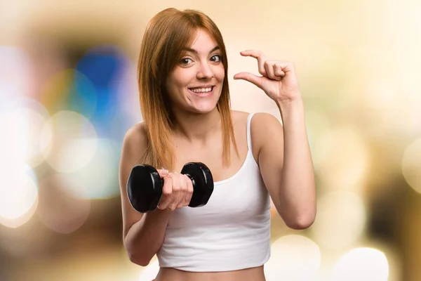 Hermosa mujer deportiva con mancuernas haciendo minúscula señal sobre fondo desenfocado — Foto de Stock