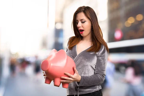 Vacker ung flicka som håller en piggybank på ofokuserat bakgrund — Stockfoto