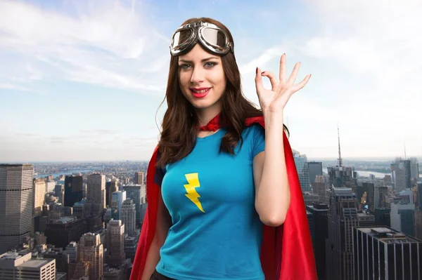 Bastante chica superhéroe haciendo OK signo con la ciudad en el fondo — Foto de Stock