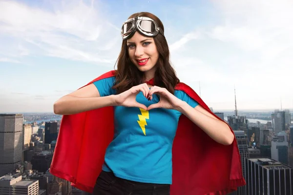 Linda chica superhéroe haciendo un corazón con sus manos con la ciudad en el fondo — Foto de Stock