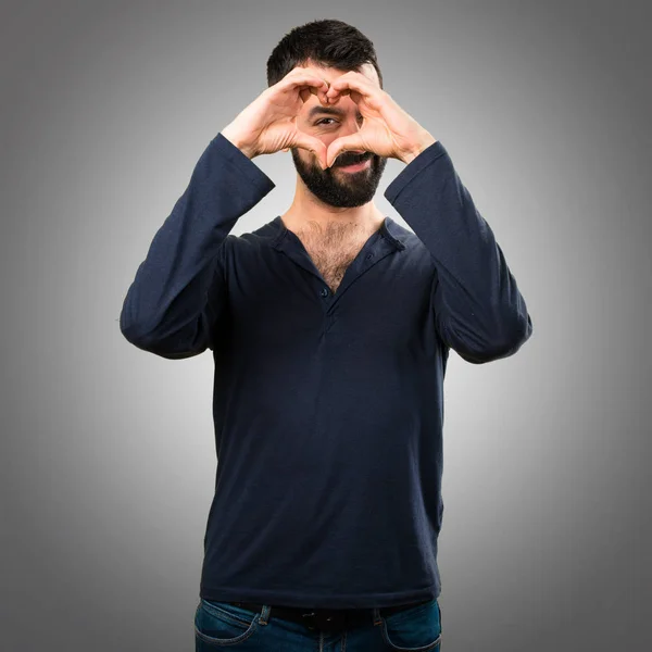 Handsome man with beard making a heart with his hands on grey ba — Stock Photo, Image
