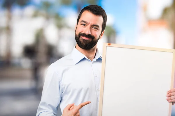 Glücklicher gutaussehender Mann mit Bart, der ein leeres Plakat über Unfokus hält — Stockfoto