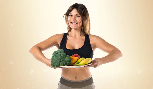 Deporte mujer sosteniendo frutas y verduras sobre fondo ocre — Foto de Stock