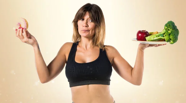 Sport woman holding fruits and vegetables in one hand and donut — Stock Photo, Image