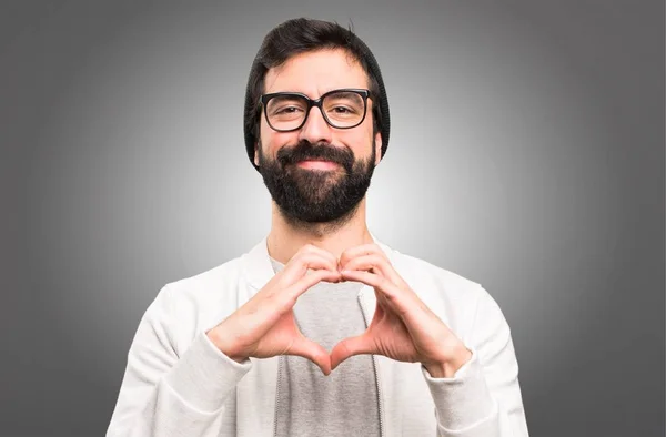 Hipster homem fazendo um coração com as mãos no fundo cinza — Fotografia de Stock