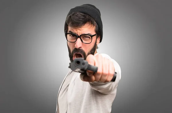 Hipster homem atirando com uma pistola no fundo cinza — Fotografia de Stock
