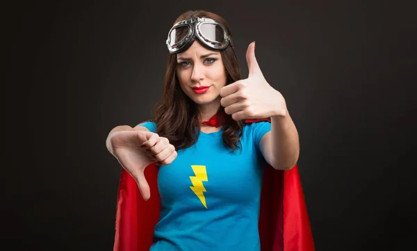 Menina super-herói bonito fazendo bom-mau sinal no fundo preto — Fotografia de Stock