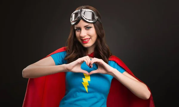 Pretty superhero girl making a heart with her hands on black background