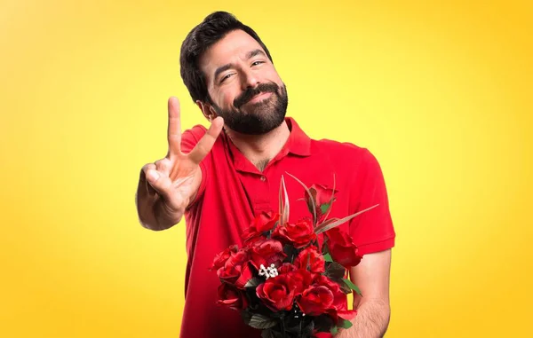 Jóképű férfi gazdaság virágok gesztus győzelmet a sárga háttér — Stock Fotó