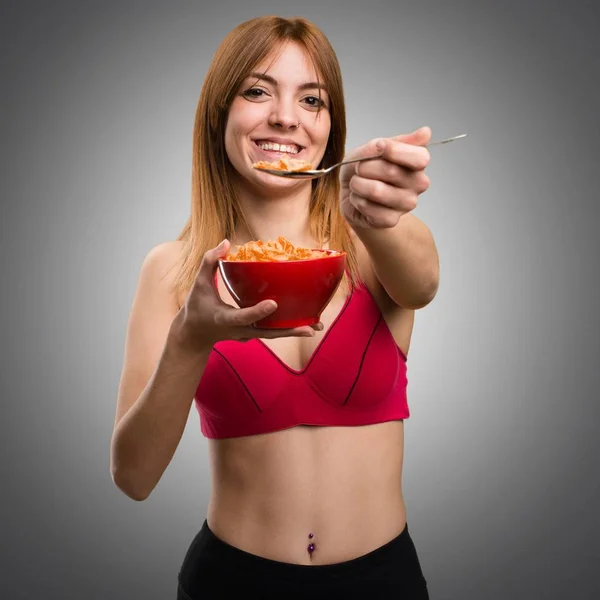 Jovem mulher esportiva comendo cereais de uma tigela em fundo cinza — Fotografia de Stock