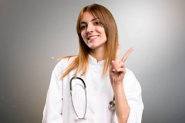 Молода лікарка робить жест перемоги на сірому фоні — стокове фото