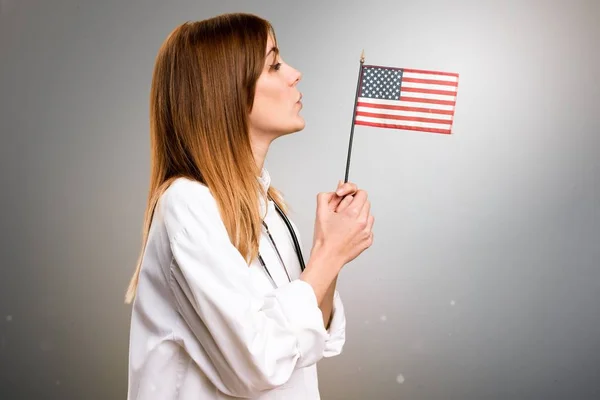 Joven doctora sosteniendo una bandera americana sobre fondo gris —  Fotos de Stock
