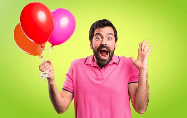 Bonito jovem segurando balões e fazendo gesto surpresa no fundo colorido — Fotografia de Stock