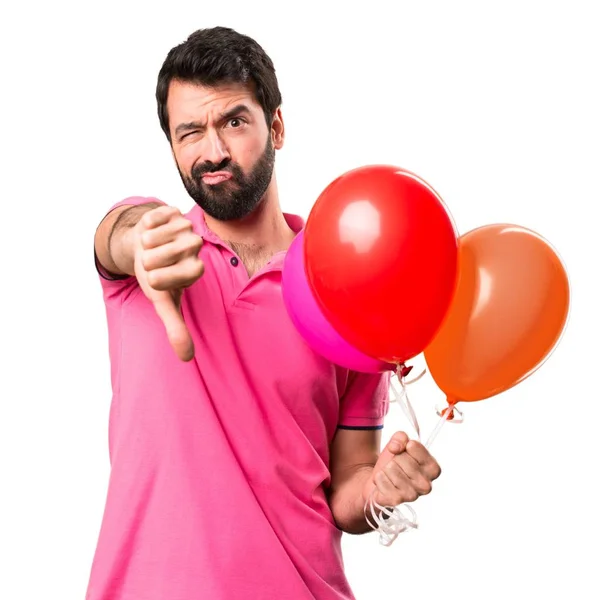 Όμορφος νεαρός άνδρας κρατά μπαλόνια και κάνοντας κακό σημάδι πάνω από το απομονωμένο λευκό φόντο — Φωτογραφία Αρχείου