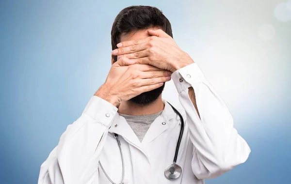 Jovem médico cobrindo seu rosto em fundo azul — Fotografia de Stock