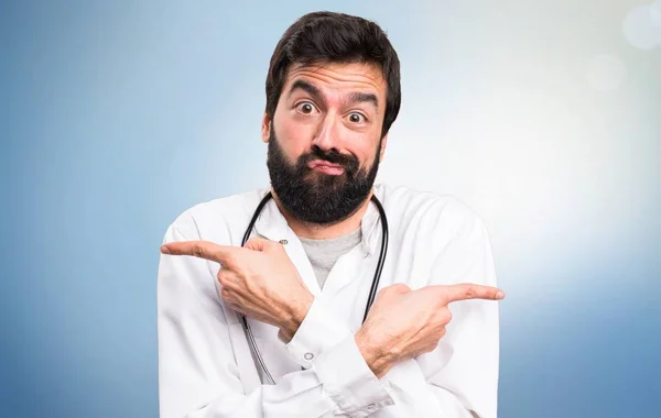 Young doctor pointing to the laterals having doubts on blue background — Stock Photo, Image