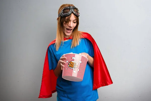 Menina super-herói bonito comer pipocas em um fundo texturizado cinza — Fotografia de Stock