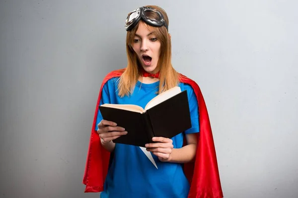 Pretty superhero girl reading book on a gray textured background — Stock Photo, Image