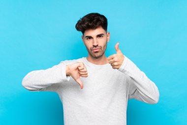 İzole edilmiş mavi arka planda genç bir adam iyiye-kötüye işaret yapıyor. Evet ya da hayır arasında kararsız