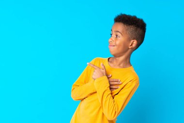 İzole edilmiş mavi arka planı olan Afro-Amerikalı çocuk bir ürünü sunmak için kenarı işaret ediyor.
