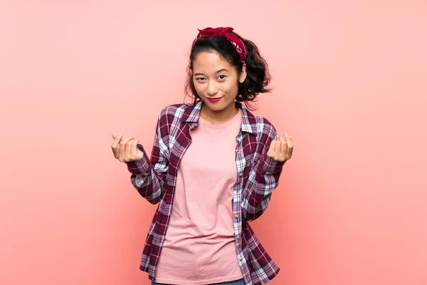 亚洲的年轻女子在孤零零的粉红背景上做着赚钱的手势 — 图库照片