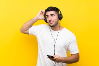 Müzik dinleyen genç yakışıklı bir adam, izole edilmiş sarı bir duvarla kuşkuları ve kafa karıştırıcı bir ifadeyle.