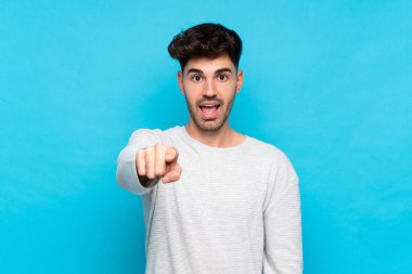 İzole edilmiş mavi arka planı olan genç adam şaşırmış ve ön tarafı işaret ediyor.