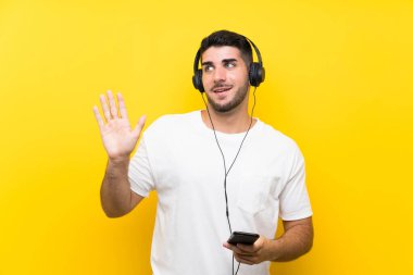 Müzik dinleyen genç yakışıklı bir adam. Seyyar, izole edilmiş sarı bir duvarla. Mutlu bir ifadeyle selam veriyor.