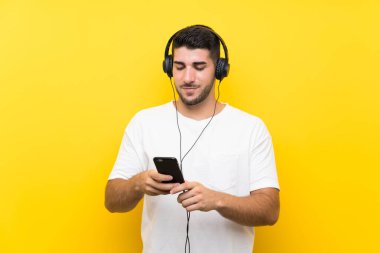 Genç yakışıklı adam izole sarı duvar üzerinde bir cep ile müzik dinliyor