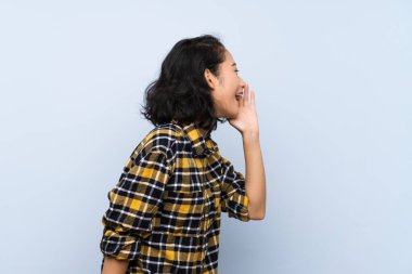 Asyalı genç bir kadın izole edilmiş mavi arka planda ağzı sonuna kadar açık bağırıyor.