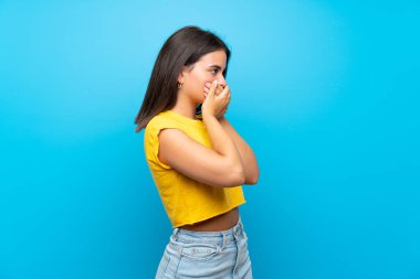 İzole edilmiş mavi arka planı olan genç bir kız ağzını kapatıp yan tarafa bakıyor.