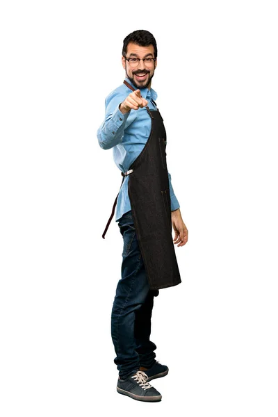 Full-length shot of Man with apron points finger at you with a confident expression over isolated white background