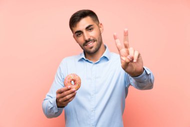 İzole edilmiş pembe arka planda donut tutan genç adam gülümsüyor ve zafer işareti gösteriyor.