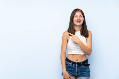 İzole edilmiş mavi arka planı olan genç bir kadın şaşırmış ve işaret ediyor.