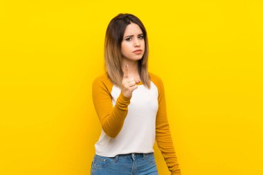 Güzel genç bir kadın, izole edilmiş sarı duvarın üzerinde hayal kırıklığına uğramış ve ön tarafı işaret ediyor.