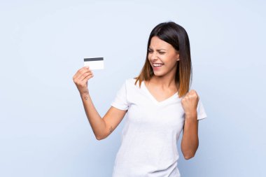 Soyutlanmış mavi arka planı olan genç bir kadın kredi kartı tutuyor.
