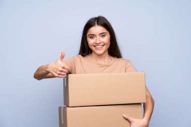 İzole edilmiş mavi arka plan üzerinde genç bir kadın bir kutuyu başparmağıyla başka bir yere taşımak için tutuyor.