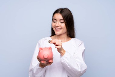 Soyutlanmış mavi arka planı olan genç bir kadın büyük bir kumbara tutuyor.