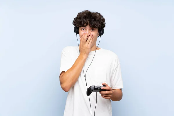 Young man playing with a video game controller over isolated blue wall with surprise facial expression