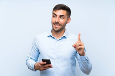 Genç ve yakışıklı bir adam cep telefonuyla mavi duvarı aşıyor. Çözümü, parmağını kaldırırken fark etmeyi umuyor.