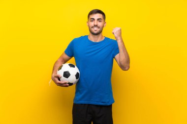 Genç yakışıklı futbolcu, izole edilmiş sarı duvarda zaferi kutluyor.